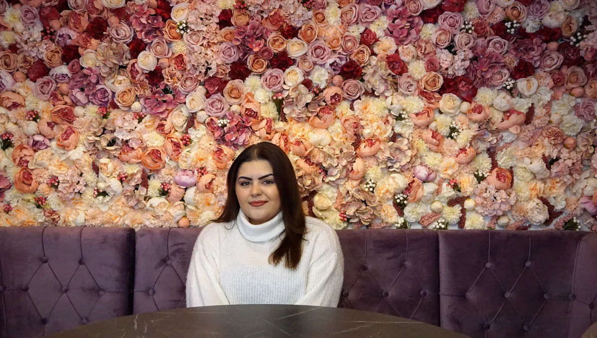 Man sieht eine Frau, die in einer Lounge sitzt. Die Wand hinter ihr ist mit Blumen dekoriert.